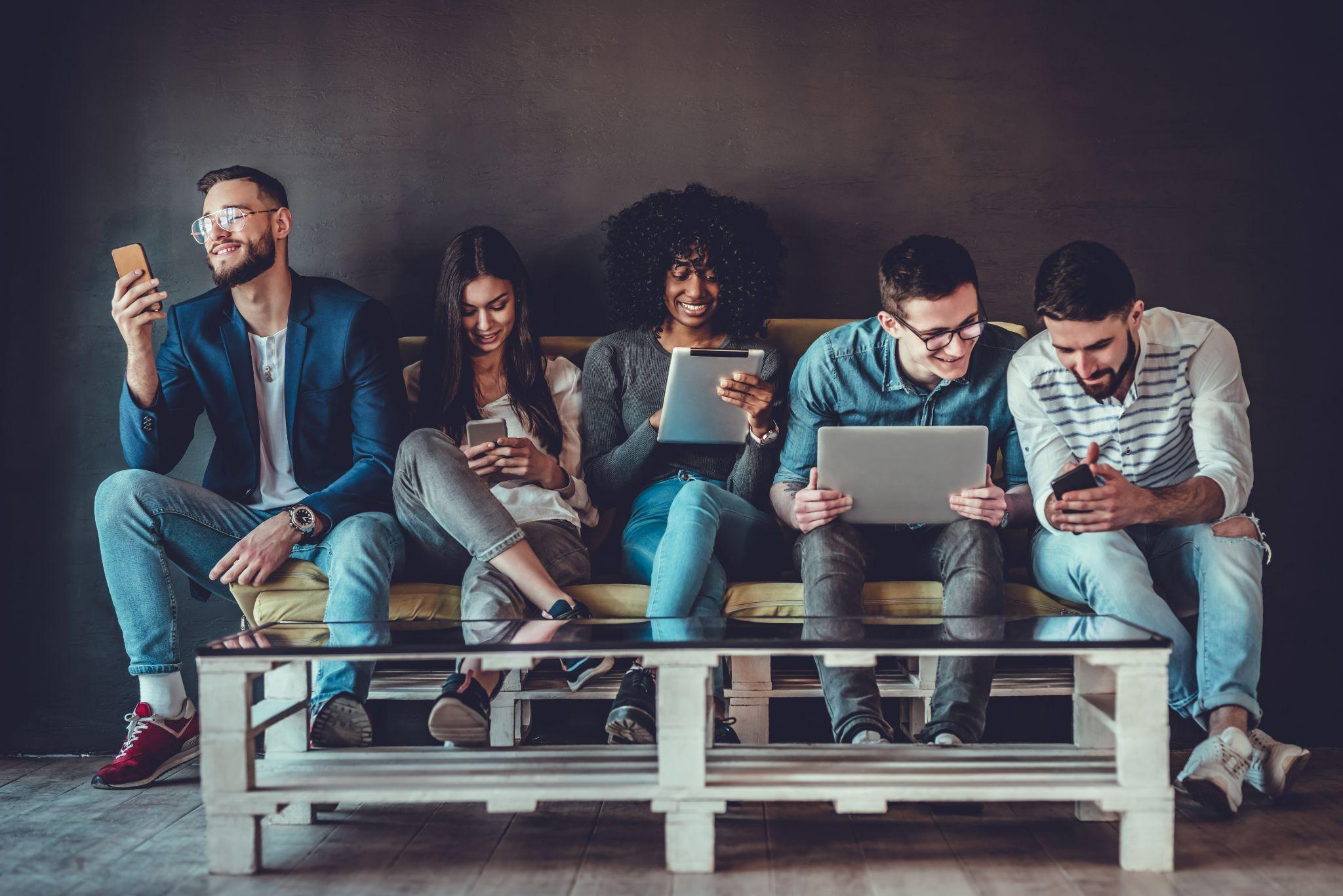 Diverse students using gadgets, sit on sofa in line on grey wall background indoors