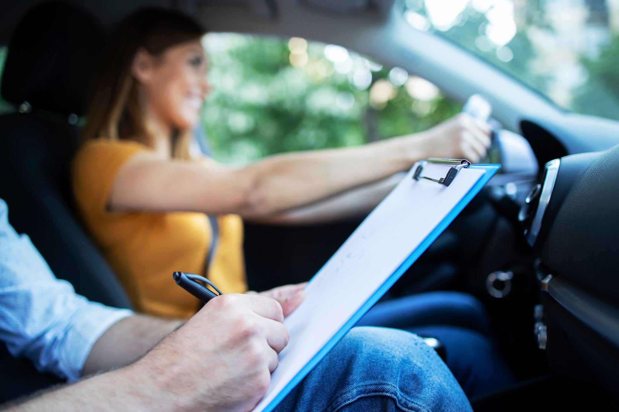 Close up view of driving instructor holding checklist while in background female student steering and driving car.