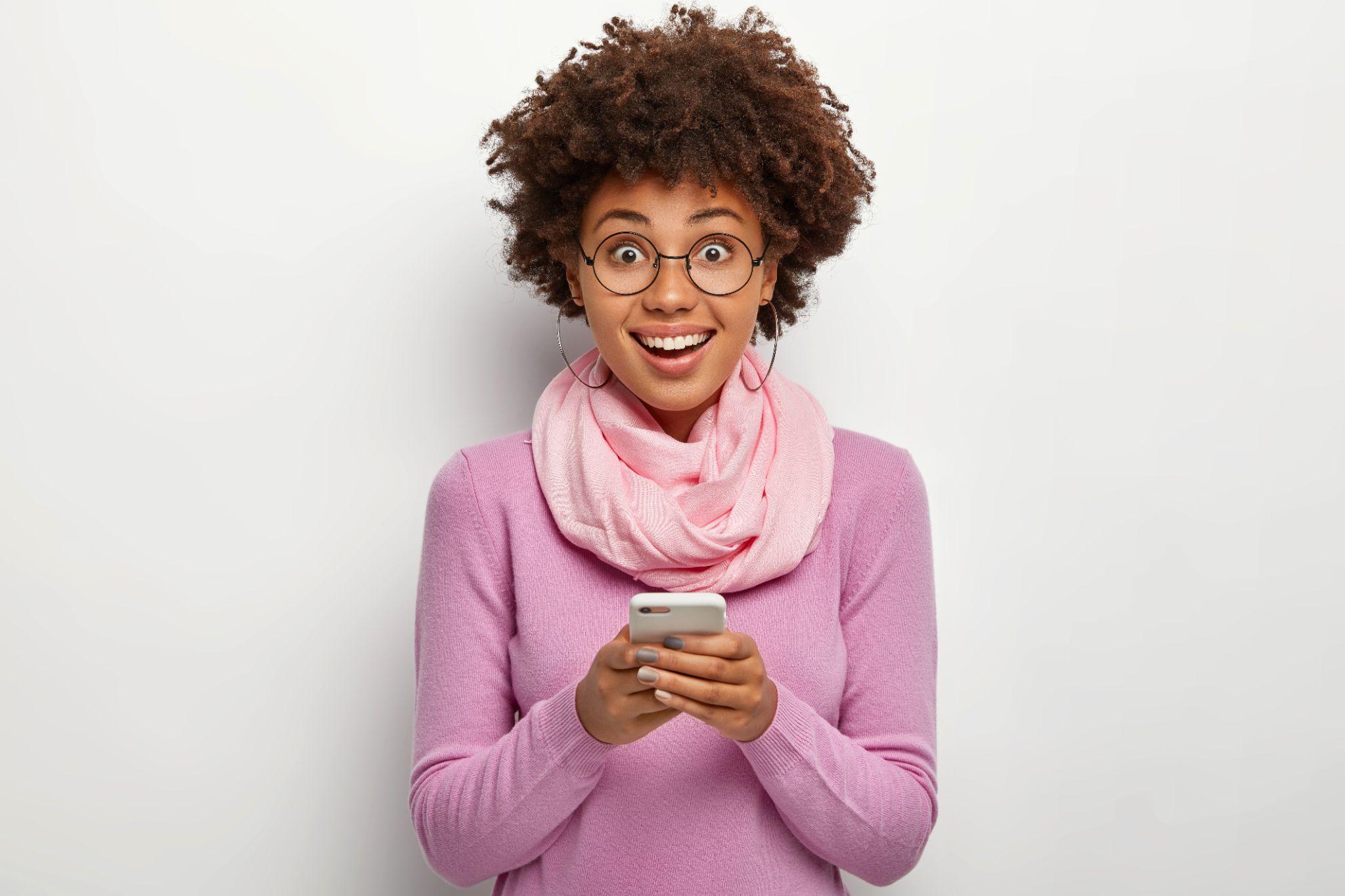 Happy curly haired woman holds modern cellular, chats online, smiles joyfully
