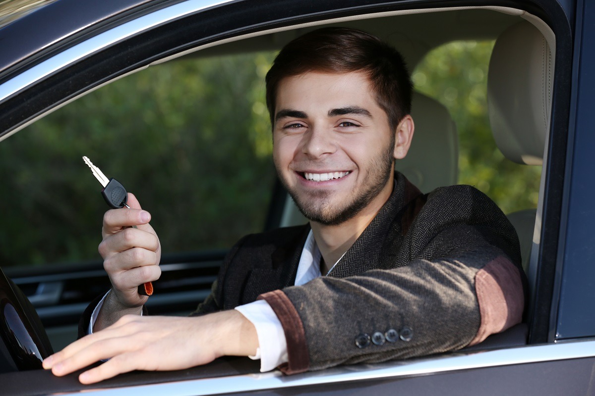 Young male driver in car holding keys out of the window and smiling.